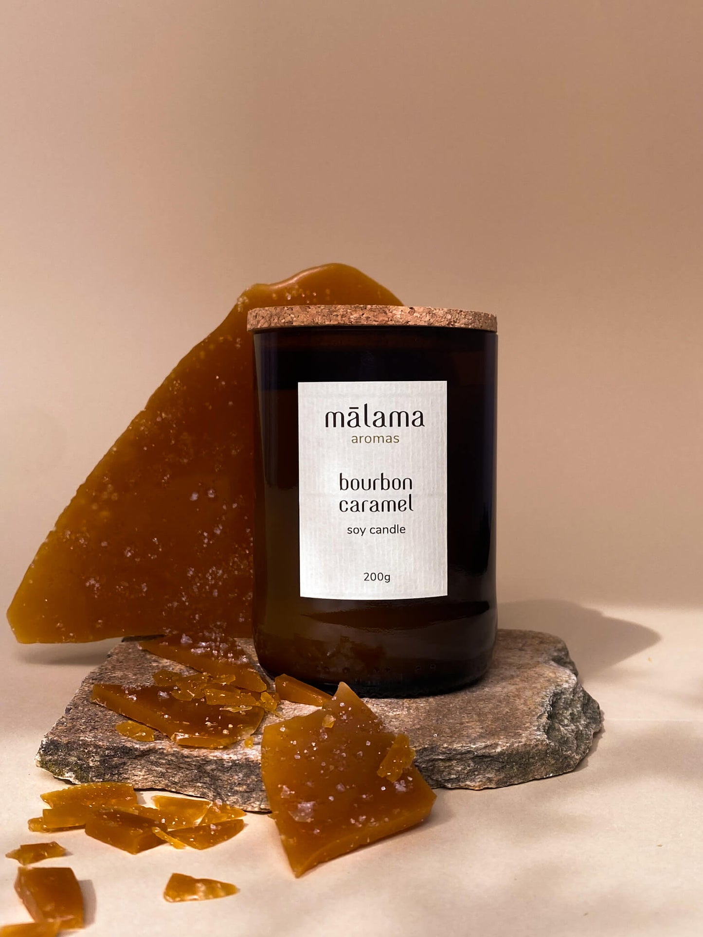 Malama Aromas bourbon caramel 200g soy candle. The candle is in a dark amber glass container with a cork lid, and it features a label that includes the brand name "malama aromas". Accompanying the candle are pieces of brittle caramel candy, displayed on a flat stone and the surrounding area, against a soft beige background. The composition suggests a warm, sweet scent and a cozy ambiance.