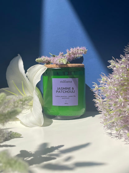 A green candle with a cork lid, labeled "mālama JASMINE & PATCHOULI," is adorned with delicate dried flowers. The label also notes the scents of "Indian jasmine, water lily, and patchouli." The candle rests on a textured stone surface against a blue background, with shadows cast by nearby light.