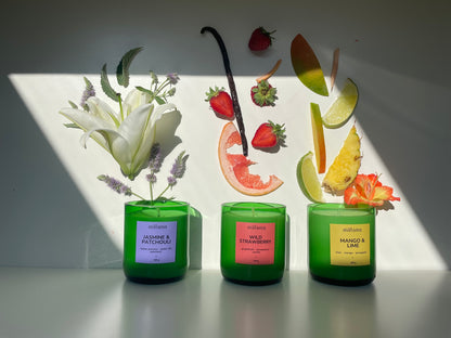 Three green glass candles with cork lids sit in sunlight. Each candle has a label and decorative topping:  "Wild Strawberry": red label, topped with a strawberry slice. "Mango & Lime": yellow label, topped with a lime wedge. "Jasmine & Patchouli": purple label, topped with delicate flowers. The shadows cast behind them add depth and contrast.