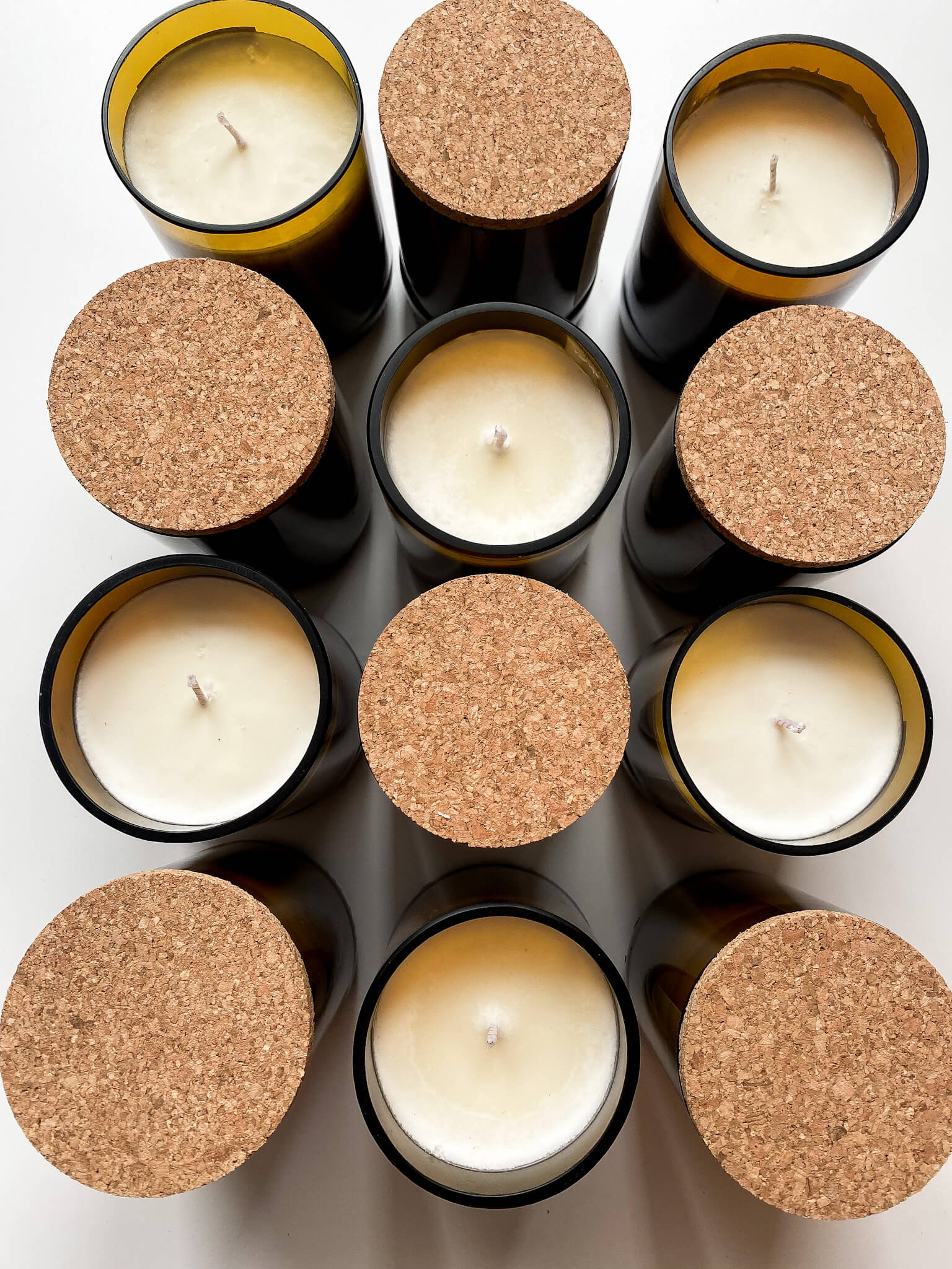 Malama Aromas unlit cylindrical candles in black and gold containers, some capped with cork lids, arranged neatly on a plain surface.