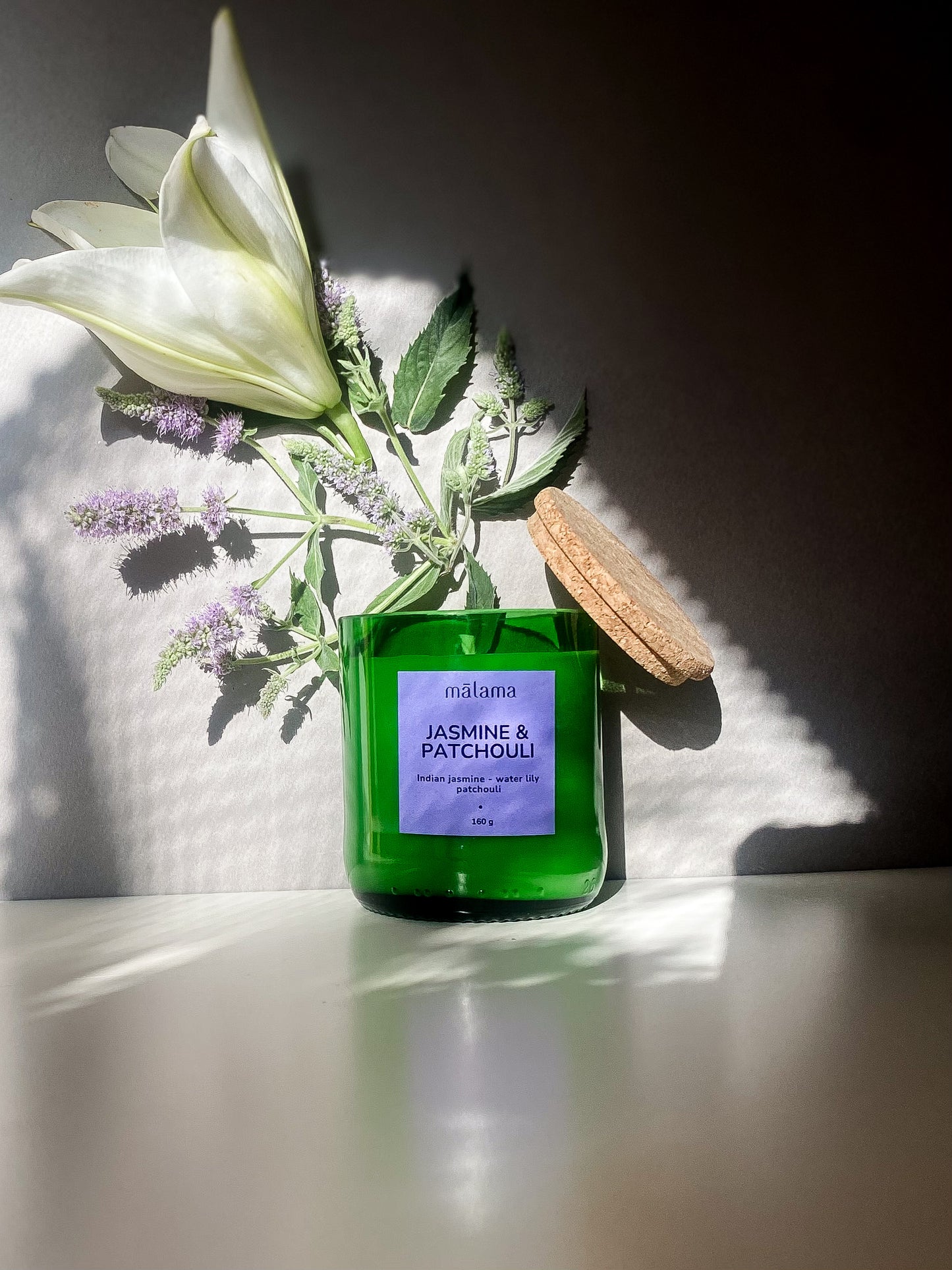 A green candle with a cork lid, labeled "mālama JASMINE & PATCHOULI," is adorned with delicate dried flowers. The label also notes the scents of "Indian jasmine, water lily, and patchouli." The candle rests on a textured stone surface against a blue background, with shadows cast by nearby light.