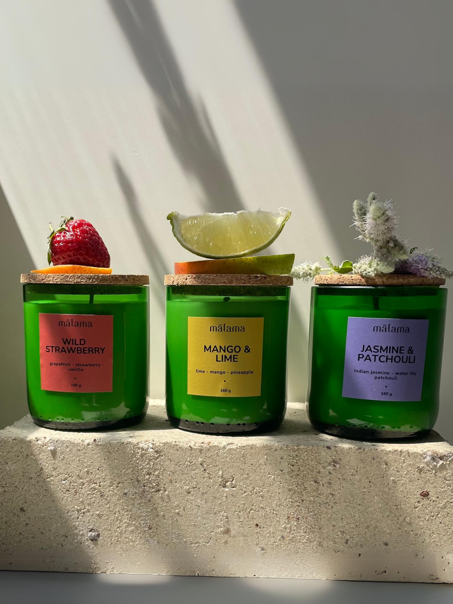 Three green glass candles with cork lids sit in sunlight. Each candle has a label and decorative topping: "Wild Strawberry": red label, topped with a strawberry slice. "Mango & Lime": yellow label, topped with a lime wedge. "Jasmine & Patchouli": purple label, topped with delicate flowers. The shadows cast behind them add depth and contrast.